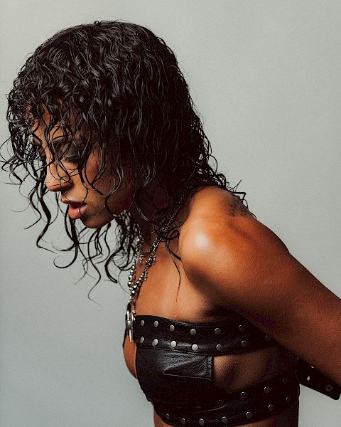 A person with wet, curly hair wearing a studded black harness-like top, side profile against a plain gray background.