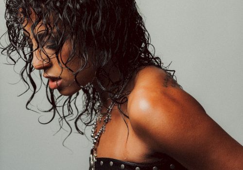 A person with wet, curly hair wearing a studded black harness-like top, side profile against a plain gray background.
