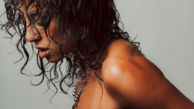 A person with wet, curly hair wearing a studded black harness-like top, side profile against a plain gray background.