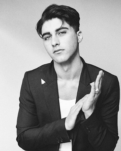 A black-and-white portrait of a stylish man in a blazer and white tee, seated, hands lightly clapping, and a confident, cool gaze.