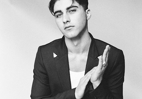 A black-and-white portrait of a stylish man in a blazer and white tee, seated, hands lightly clapping, and a confident, cool gaze.