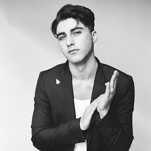 A black-and-white portrait of a stylish man in a blazer and white tee, seated, hands lightly clapping, and a confident, cool gaze.