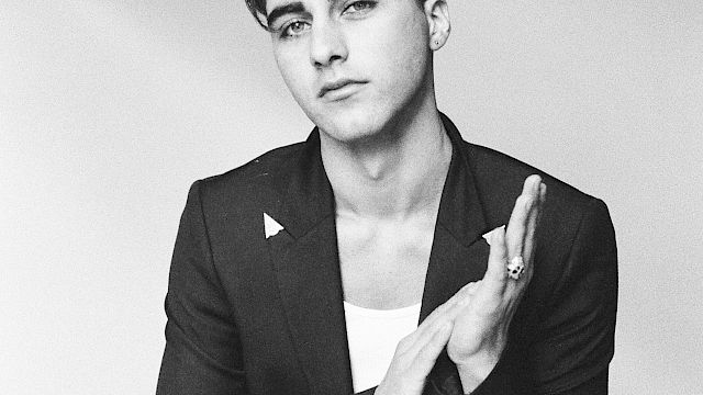 A black-and-white portrait of a stylish man in a blazer and white tee, seated, hands lightly clapping, and a confident, cool gaze.
