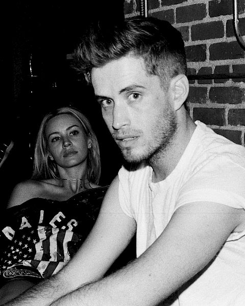 Two people sit in a dim, brick-walled room; a man in a white T-shirt gazes at the camera while a woman sits behind him, looking on.