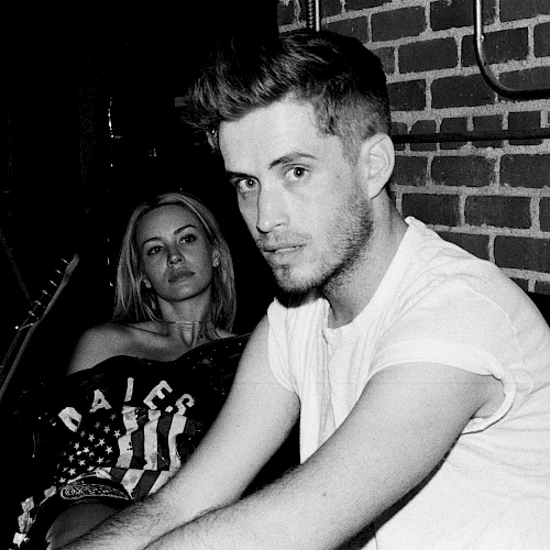 Two people sit in a dim, brick-walled room; a man in a white T-shirt gazes at the camera while a woman sits behind him, looking on.