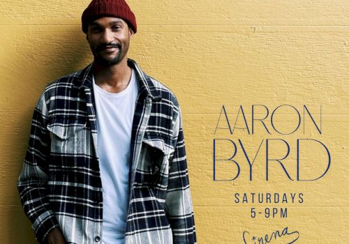 A stylish man in a red beanie and plaid shirt stands against a yellow wall with &ldquo;ARON BYRD&rdquo; promotional text.