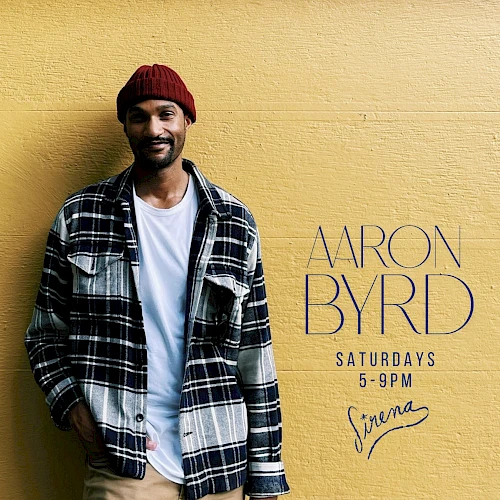 A stylish man in a red beanie and plaid shirt stands against a yellow wall with &ldquo;ARON BYRD&rdquo; promotional text.