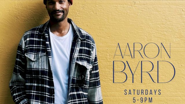 A stylish man in a red beanie and plaid shirt stands against a yellow wall with &ldquo;ARON BYRD&rdquo; promotional text.