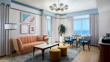 Bright living room with blue accents, sofa and coffee table, dining set by ocean-view windows, chandelier overhead.