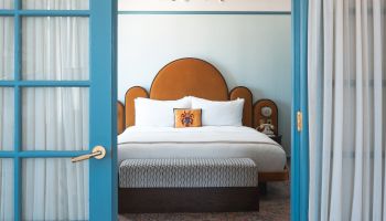 View into a stylish hotel bedroom with a blue door, king bed, chandelier, and sheer curtains framing the windows.