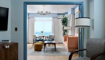 Bright coastal living room with blue trim, chandelier, curtains, ocean view, sofa, and dining table.