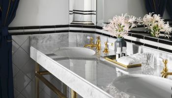 A luxurious bathroom scene with a marble sink, gold faucets, soap bottles, and pink-white flowers on a tray by the window.