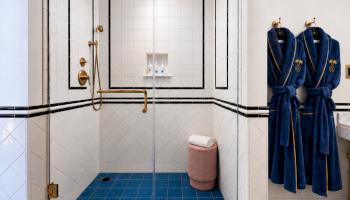 A luxury bathroom shower with gold fixtures, blue floor tiles, a folded towel, and two blue bathrobes hanging.