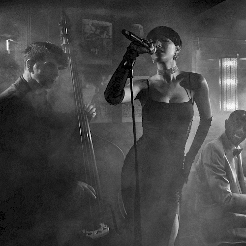 A moody black-and-white jazz trio: a singer at center with a microphone, a pianist on the right, and a guitarist/bassist on the left in smoky ambiance.