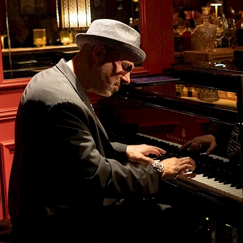 A man in a hat and sunglasses plays a piano in a dimly lit club, konzentred on the keys.