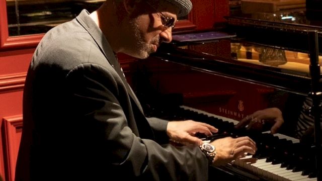 A man in a hat and sunglasses plays a piano in a dimly lit club, konzentred on the keys.