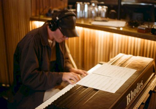 A musician plays a Rhodes electric piano, reading sheet music at a keyboard in a dim room.