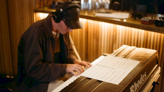 A musician plays a Rhodes electric piano, reading sheet music at a keyboard in a dim room.