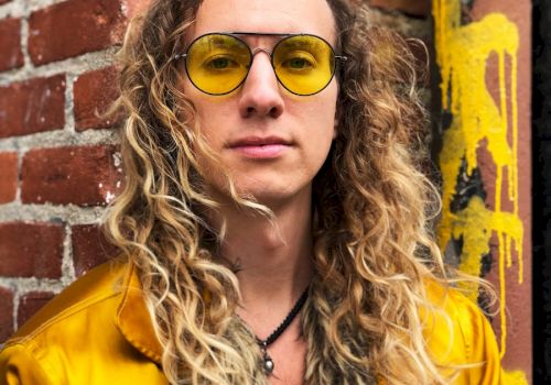 Woman with curly hair in a yellow jacket and round yellow sunglasses, standing in front of a brick wall with graffiti.