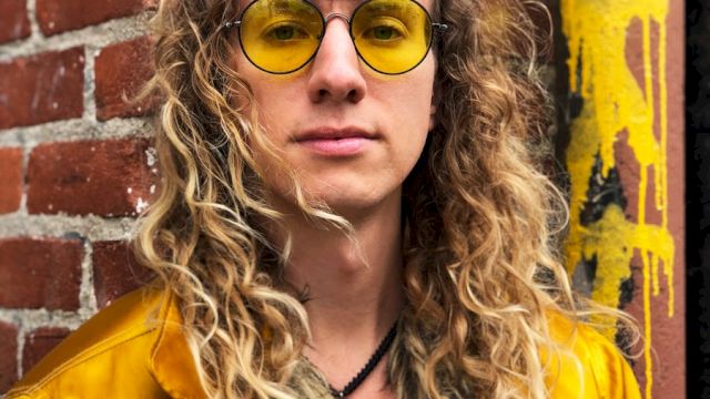 Woman with curly hair in a yellow jacket and round yellow sunglasses, standing in front of a brick wall with graffiti.
