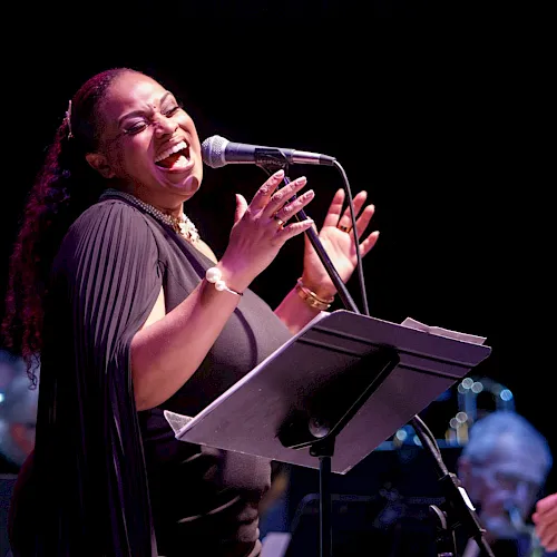 A singer performs onstage at a microphone, smiling and singing from sheet music while a band plays in the background.