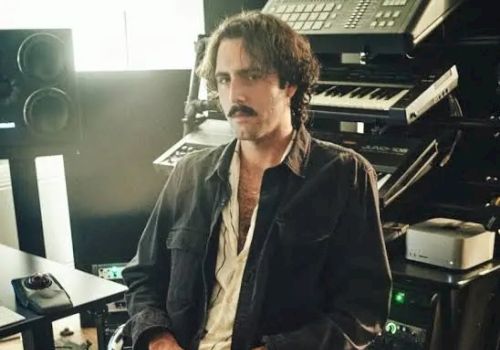 A man with a mustache sits at a desk in front of large old computer equipment, looking toward the camera.