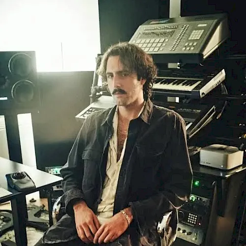 A man with a mustache sits at a desk in front of large old computer equipment, looking toward the camera.