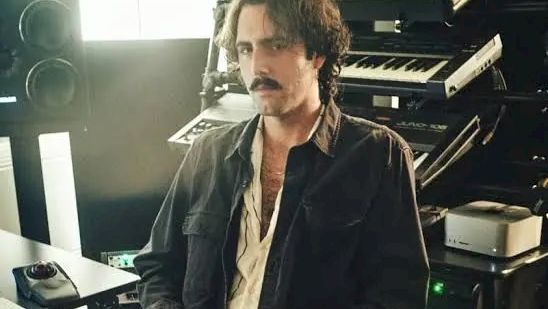 A man with a mustache sits at a desk in front of large old computer equipment, looking toward the camera.