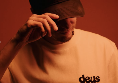 A man in a black cap and &ldquo;deus records&rdquo; T-shirt tips his hat, posing against a red backdrop.