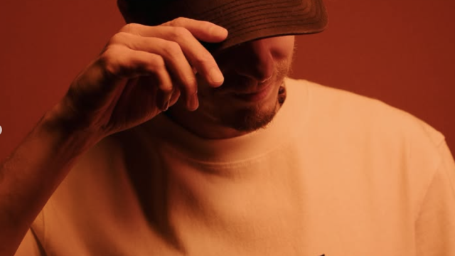 A man in a black cap and &ldquo;deus records&rdquo; T-shirt tips his hat, posing against a red backdrop.
