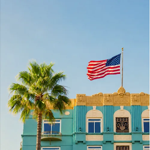A tall palm tree and a turquoise building sit beneath a waving United States flag flying from a rooftop pole.