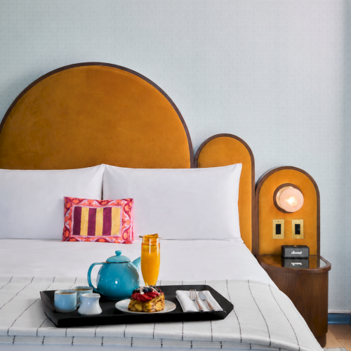 A neatly made hotel bed with colorful pillows and a serving tray of tea set, juice, and pastries on the sheets.