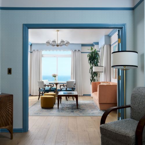 A bright seaside living room with blue trim, a chandelier, curtains, and a dining table framed by an open doorway.
