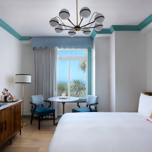 Bright hotel room with a modern chandelier, teal trim, large window, table/chairs, and a neatly made bed with a sea view.