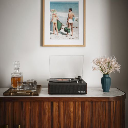 A vinyl record player sits on a table beside whiskey, glasses, and flowers, with a framed beach photo on the wall above.