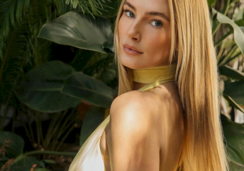 A blonde woman with bare shoulders poses among large green tropical leaves in warm sunlight.