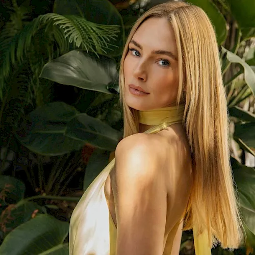 A blonde woman with bare shoulders poses among large green tropical leaves in warm sunlight.