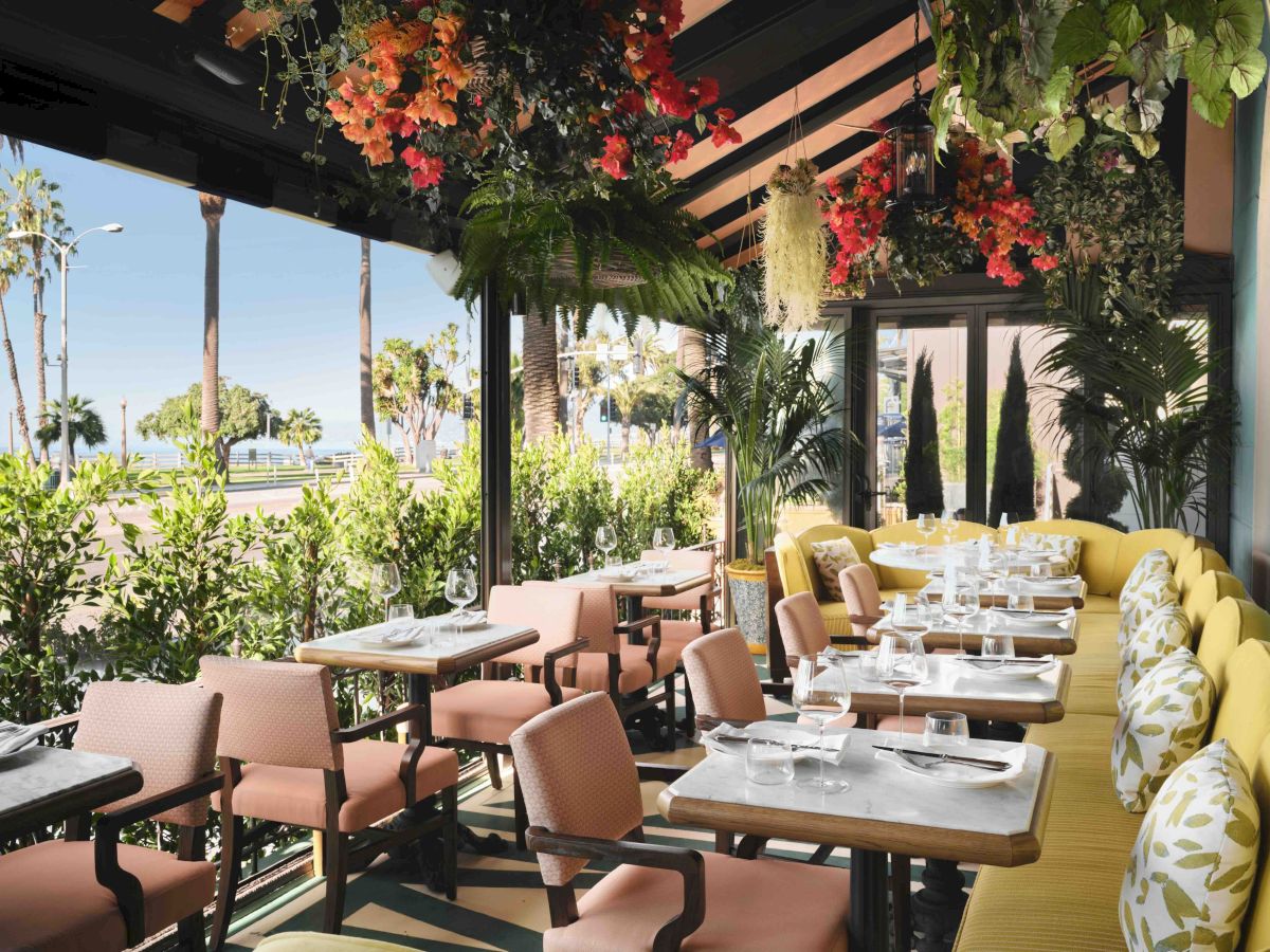 Outdoor restaurant patio with tables, glassware, and lush hanging flowers and greenery by the seaside.
