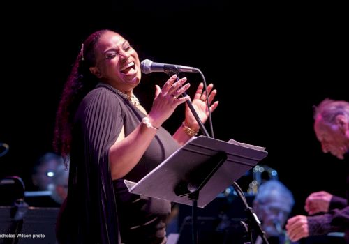 A singer performs onstage at a microphone, smiling and singing from sheet music while a band plays in the background.