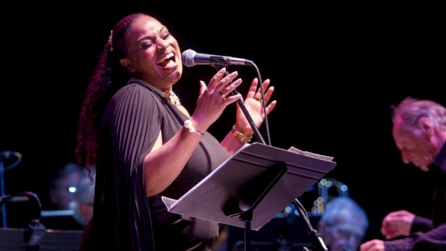 A singer performs onstage at a microphone, smiling and singing from sheet music while a band plays in the background.
