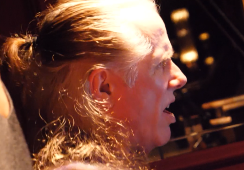 A man with long gray hair tied back sits in a dim room, eyes closed, lips parted as someone holds his shoulder in a supportive grip.