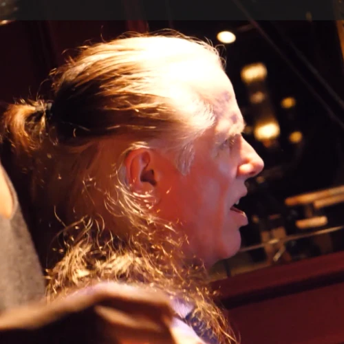 A man with long gray hair tied back sits in a dim room, eyes closed, lips parted as someone holds his shoulder in a supportive grip.