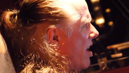 A man with long gray hair tied back sits in a dim room, eyes closed, lips parted as someone holds his shoulder in a supportive grip.