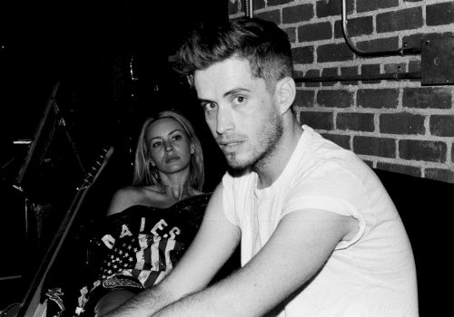 Two people sit in a dim, brick-walled room; a man in a white T-shirt gazes at the camera while a woman sits behind him, looking on.