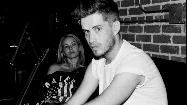 Two people sit in a dim, brick-walled room; a man in a white T-shirt gazes at the camera while a woman sits behind him, looking on.