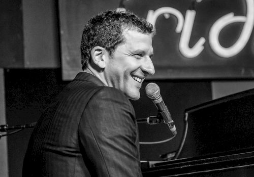 A smiling pianist singing into a mic at a piano, performing on stage.