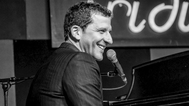 A smiling pianist singing into a mic at a piano, performing on stage.