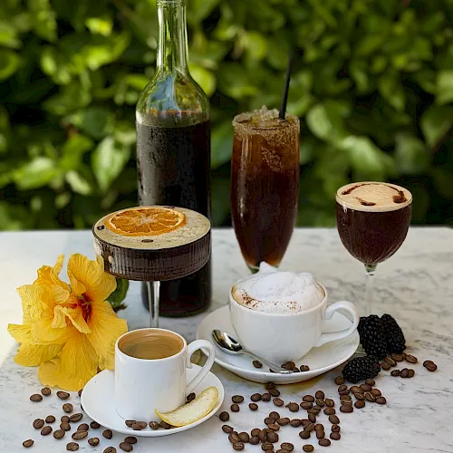 Coffee-themed setup: mugs with foamy coffee, an iced drink, coffee beans, and two bottles of dark and green liquid on a table.