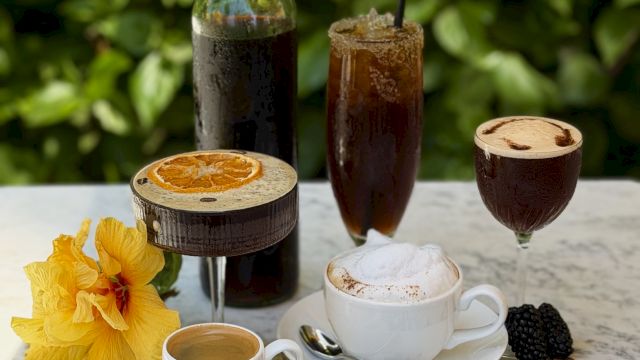 Coffee-themed setup: mugs with foamy coffee, an iced drink, coffee beans, and two bottles of dark and green liquid on a table.