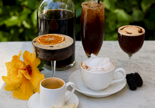 A sunny drink setup: a bottle of dark syrup, iced tea/coffee in glasses, espresso, lemon slice, blackberries, and yellow flowers.